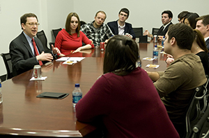 Michael Schill sits a conference table in a suit and tie. Next to him is a woman in a red dress. Ten others sit around the table with cookies and soda cans in front of them.