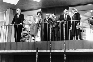 Four men and two women stand on a balcony where ribbons trail from the railing.