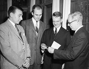 Four white men in suits and ties look at piece of paper.