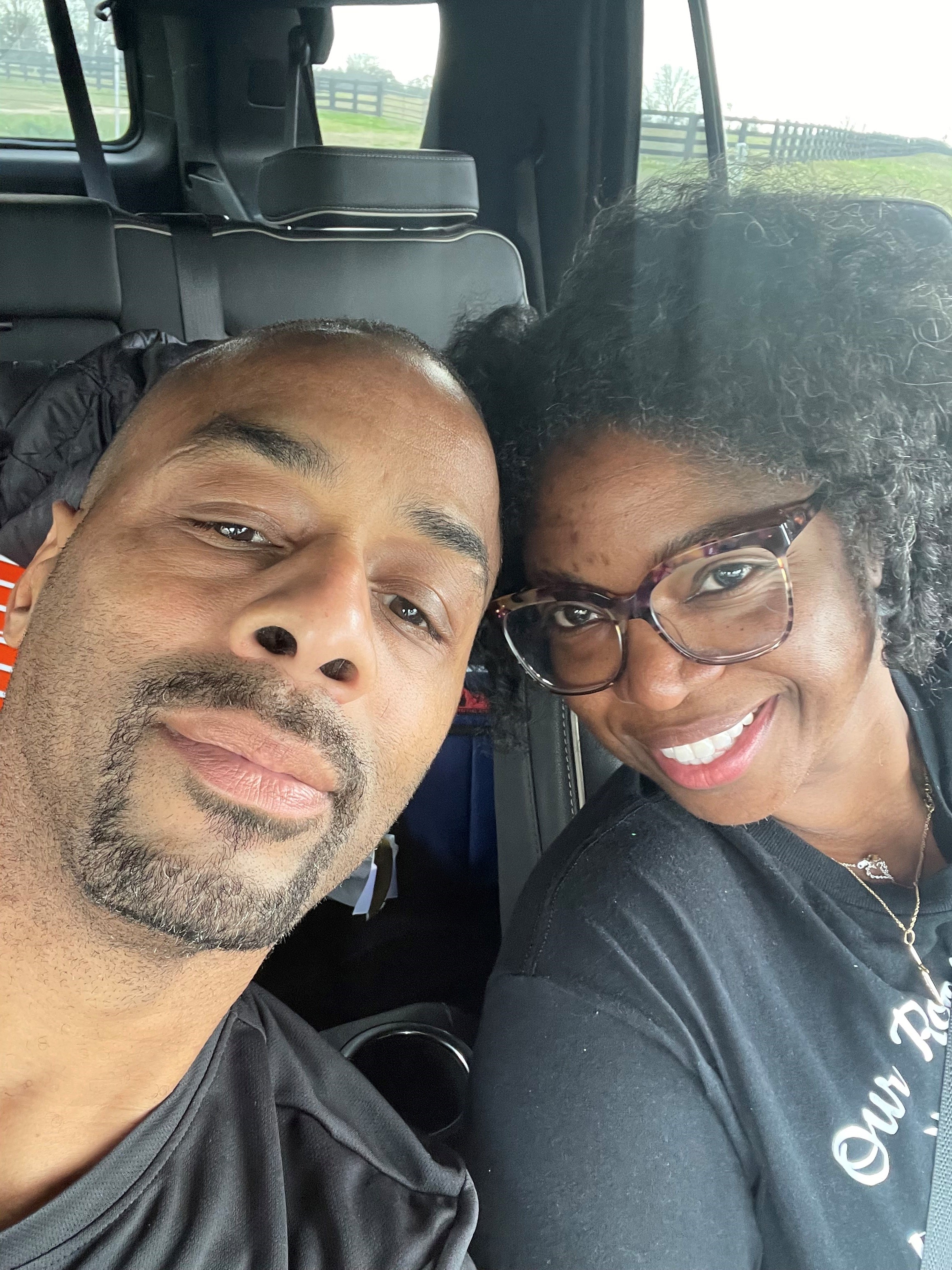 A man and a woman sitting in a car and resting their heads against one another while looking at the camera.