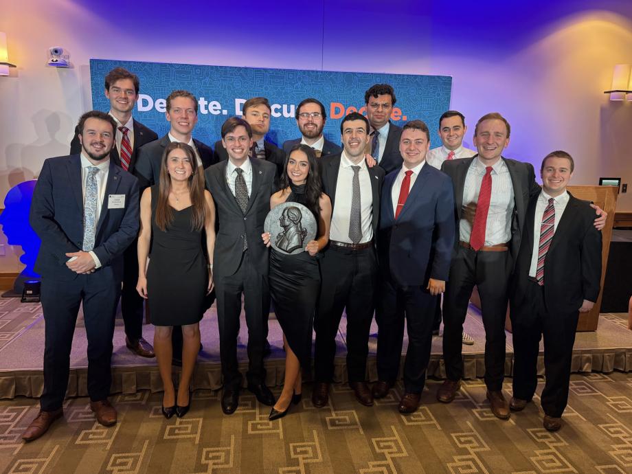 A group of students pose together for a photograph with a woman in the center holding an award plaque.