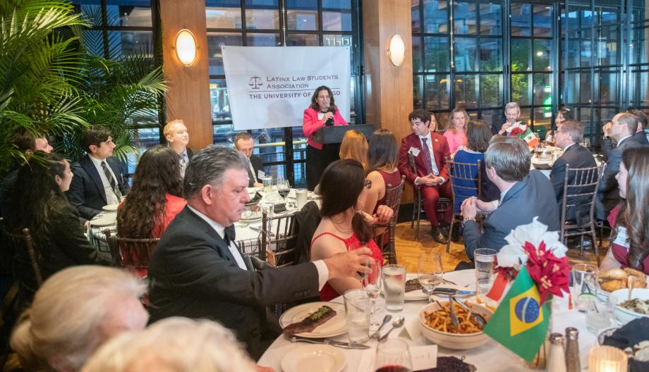 Woman standing at a lecturn addressing a room filled with people who are sitting at tables.