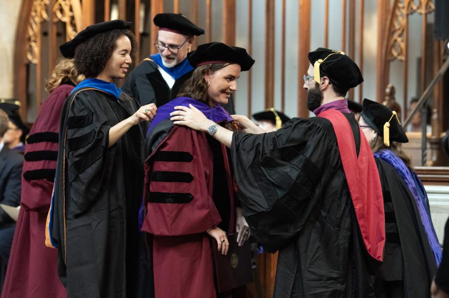 On stage, a student is hooded by Farah Peterson and John Rappaport.