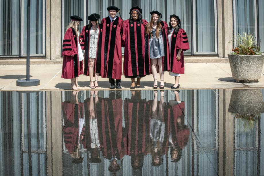 Six students, in caps and gowns, prepare for a posed photo with their reflection in the water below.