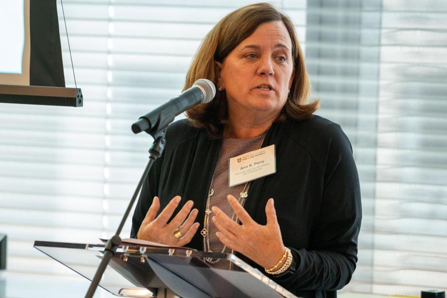 Ann Perry speaking at a microphoned podium gesturing with her hands facing upwards while talking about the Rubenstein Scholars program