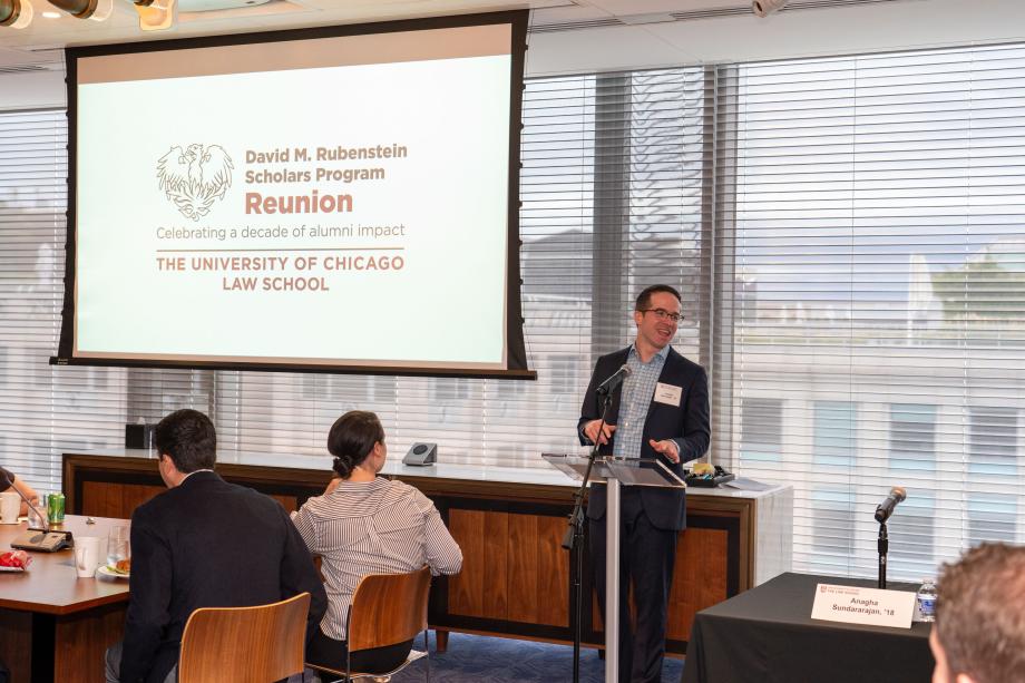 Joe Schroeder, wearing a blue suit and open collar checkered shirt, stands at a podium to introduce the event. The display behind him says, “David M. Rubenstein Scholars Program Reunion. Celebrating a decade of alumni impact. The University of Chicago law School.