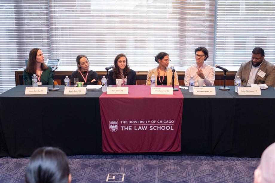 Five panelists sit a table draped with a University of Chicago Law School tablecloth. One panelist, wearing glasses and a white button down shirt, speaks into the tabletop microphone while the other panelists look on.