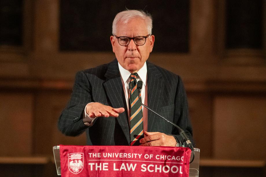 David Rubenstein, wearing a pinstripe suit and striped tie, speaks at a podium draped with a University of Chicago Law School flag. He gestures, with his palm facing downward.