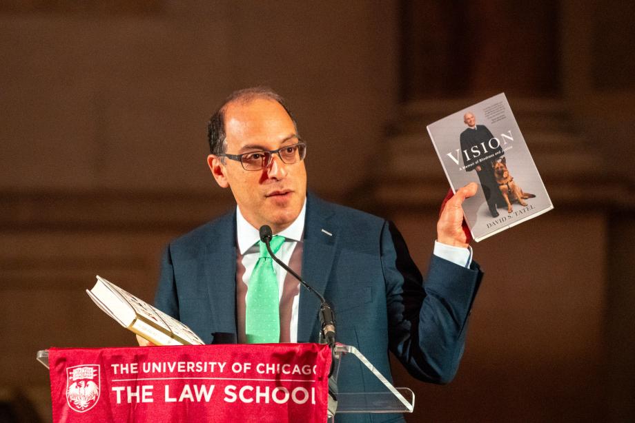 Lior Strahilevitz standing at a podium draped with a flag showing the UChicago Law logo. Strahilevitz speaks into the microphone while holding up a copy of Judge David Tatel's book, Visions, featuring David Tatel on the cover with his service animal.