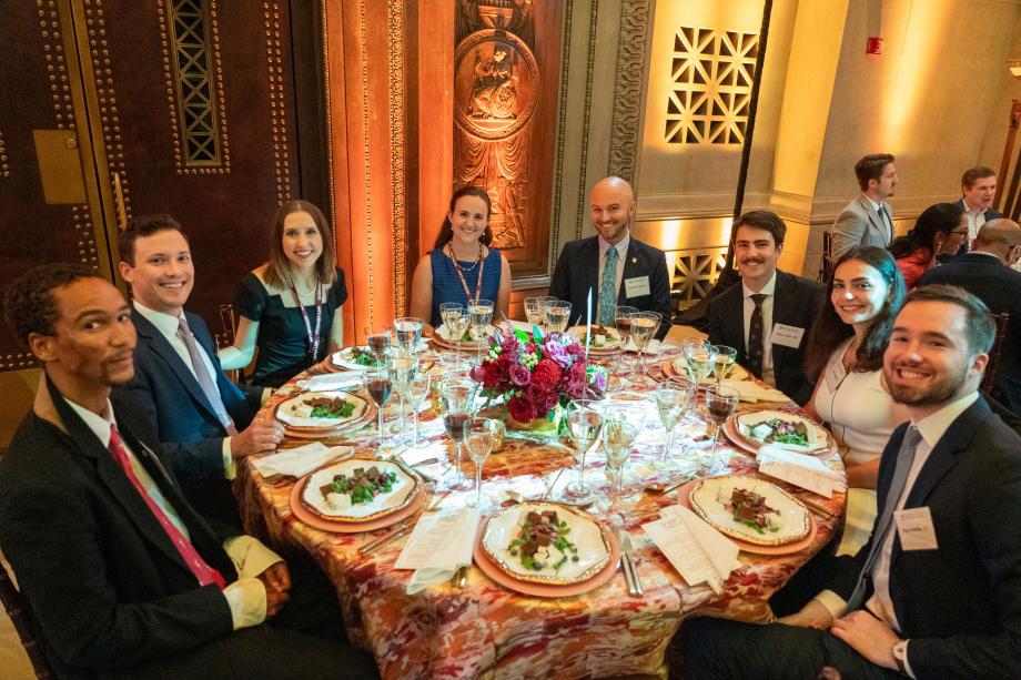 Event attendees sit around a circular table with plates of salad in front of them. All of the attendees at the table smile at the camera.