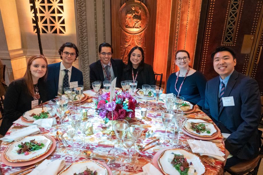 Event attendees sit around a circular table with plates of salad in front of them. All of the attendees at the table smile at the camera.