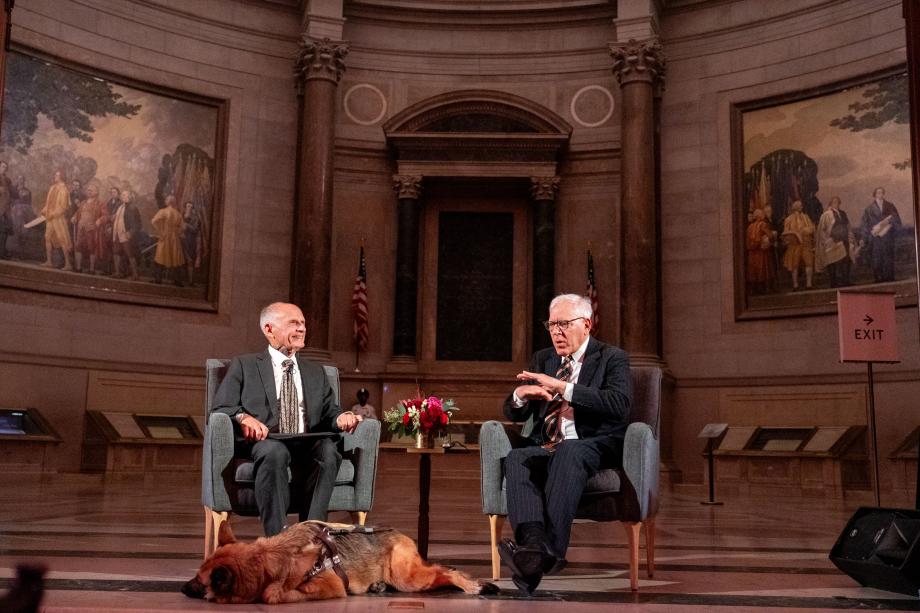 Judge David Tatel sits with Rubenstein in individual chairs in front of the audience of attendees. Tatel smiles at Rubenstein while Rubenstein speaks, and Tatel's service animal lays on the floor in front of him.