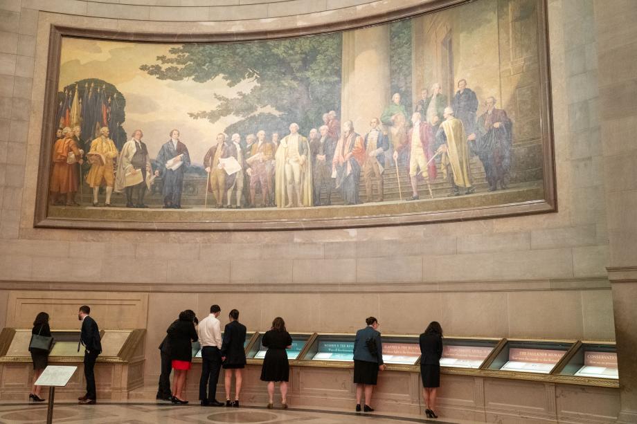 An enormous realistic painting of Revolutionary era American men is on the wall. Below the painting, a group of people look at documents behind glass cases.