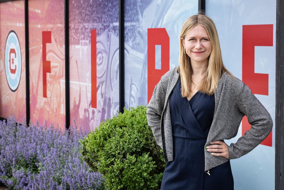 Shelby Klose, wearing a blue dress and grey cardigan, stands in front of windows with the Chicago Fire logo.