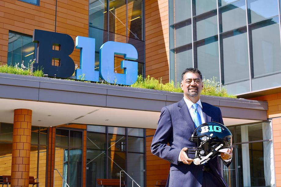 Anil Gollahalli, in a pinstripe suit, stands in front of a building with the Big Ten logo on the front. He holds a football helmet that also displays the Big Ten logo.