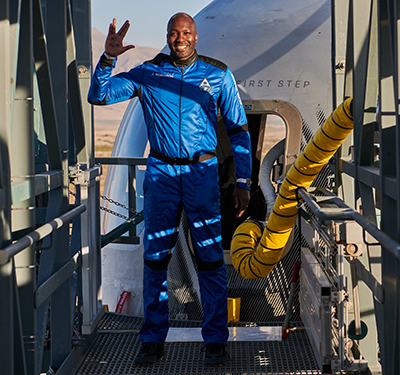 In a blue jumpsuit in front of the entrance bridge to a rocket cockpit, Jaison Robinson makes a Star Trek sign.