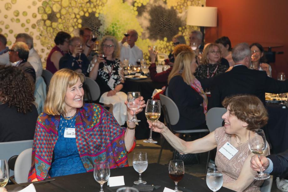 Two smiling women holding up stem glasses and offering a cheers.