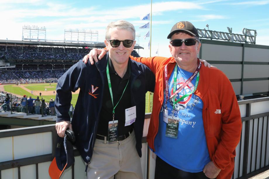 Two men stand with their arms on each other's shoulders smiling at the camera on the rooftop of a baseball field, on a bright sunny day.