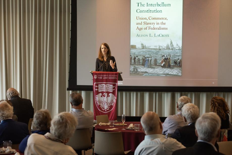 A woman standing at a lectern addressing a room full of people with a projector screen behind her.