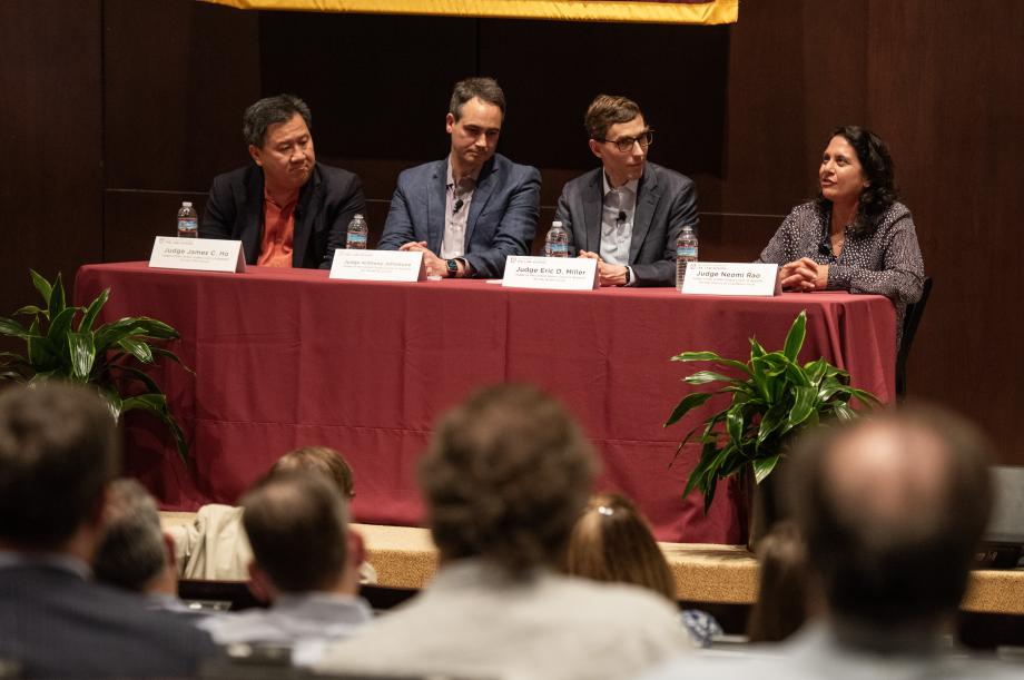 A panel of three men and one woman address a seated audience in an auditorium.