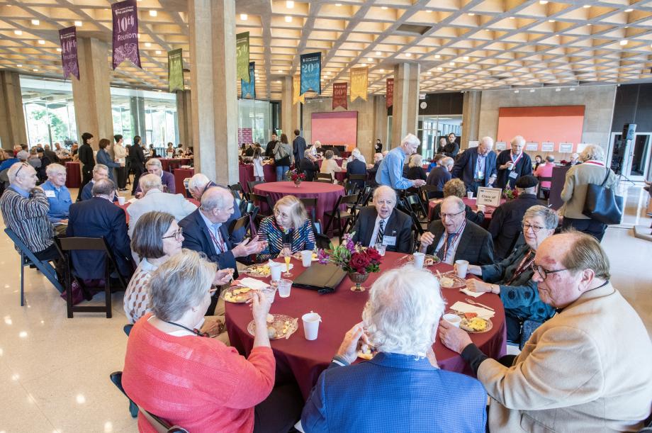 People seated at round tables, eating, in a large open room.