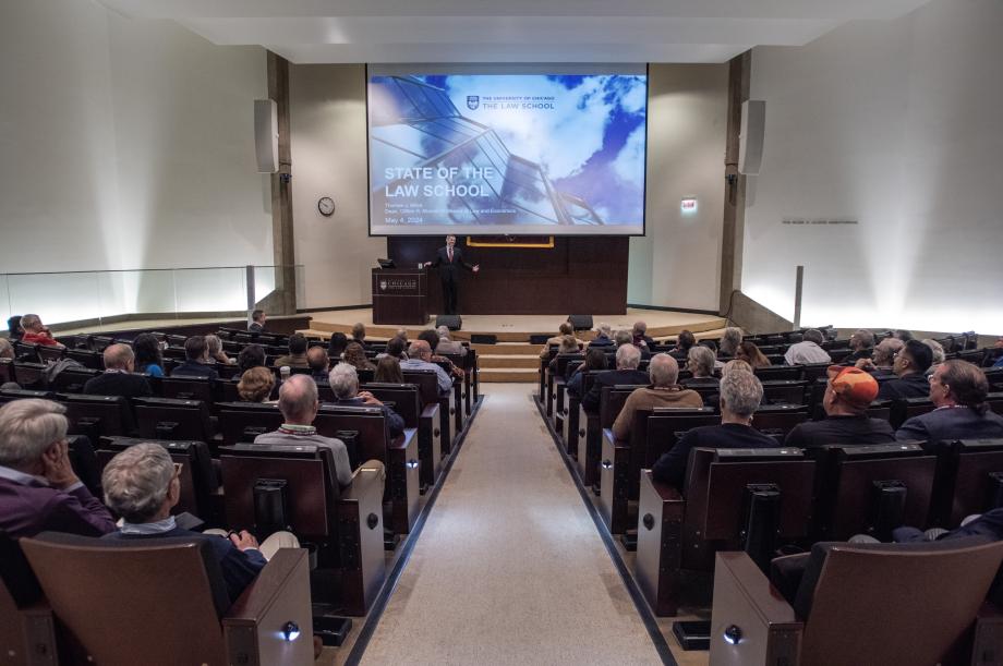 An auditorium filled with a seated audience listening to a man standing near a podium with a screen behind him.