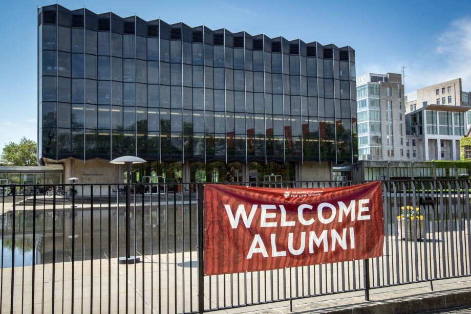 A large rectangular glass building stands tall against a bright blue sky. A banner hangs on a gate in front of it that says Welcome Alumni.