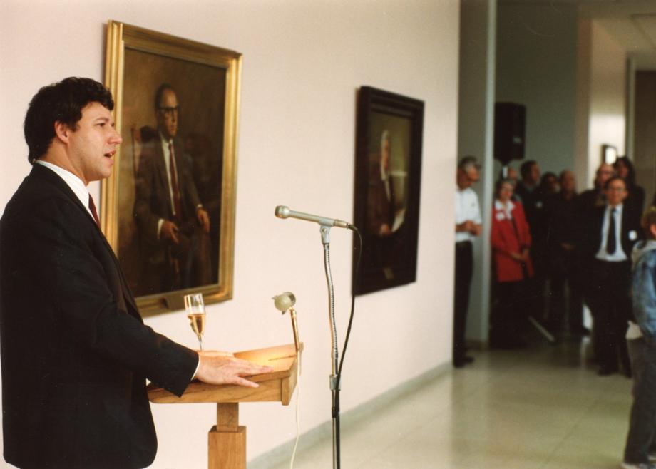 Geoffrey Stone at the Law School Dean’s Lunch, 1990. Stone, at a lectern in a hallway with portraits on the wall and people in the background look on.