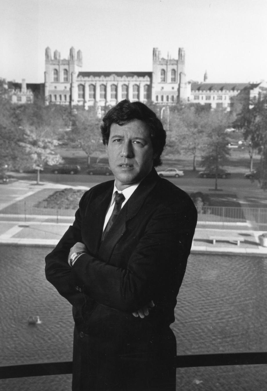 Stone in suit stares at the camera with campus buildings behind him.