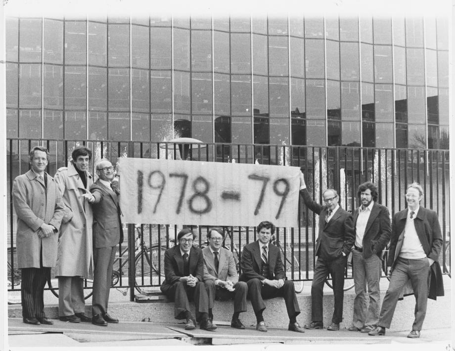 Nine men in suit jackets stand in front of the Law School building, holding a banner that says, "1978-79"