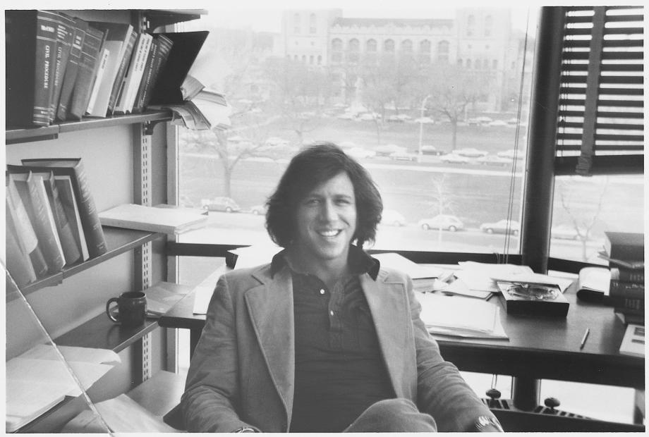 Stone, in a polo and jacket, sits at his desk with campus views behind him.