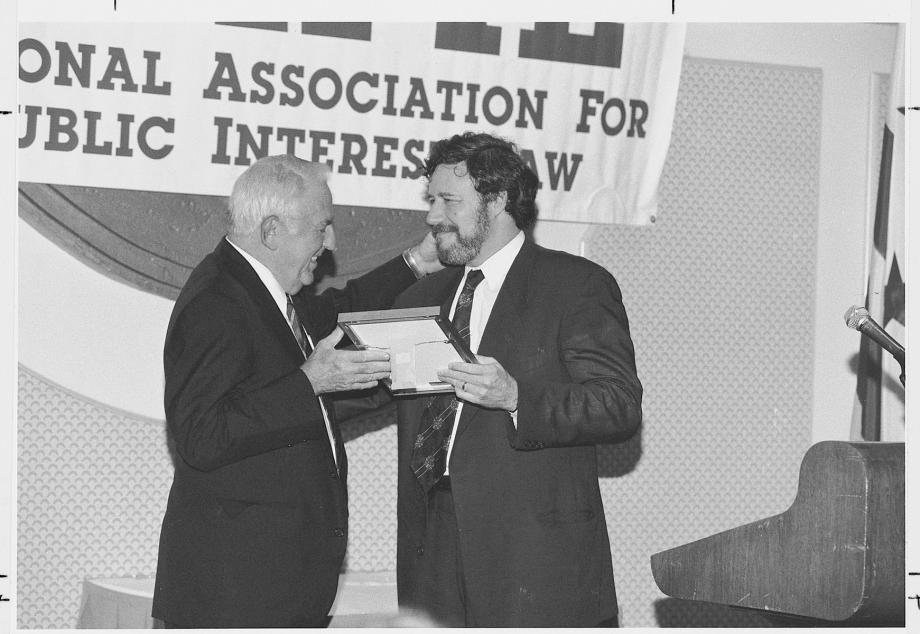 The late Abner Mikva, ’51, presents Geoffrey Stone with the National Association for Public Service Award, circa 2000. Stone, in a suit and tie, accepts a framed award from Mkiva, in a suit and tie, on a stage with a banner behind them.