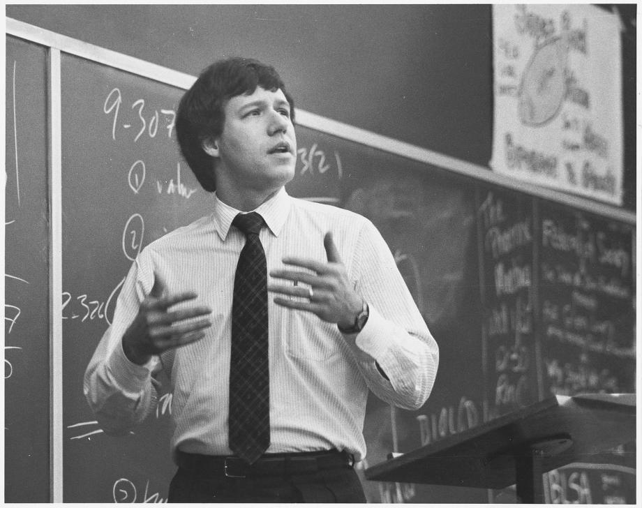 Stone, in a shirt and tie, motions with his hands at the front of a classroom with a blackboard behind him.