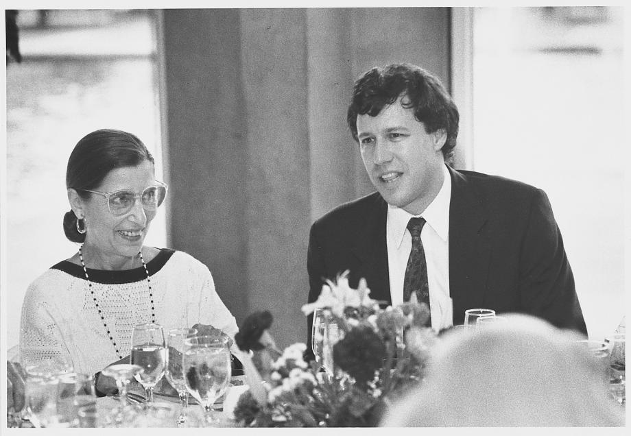 Stone, in a suit and tie, sits next to Bader Ginsburg, in a dress, who smiles at him.