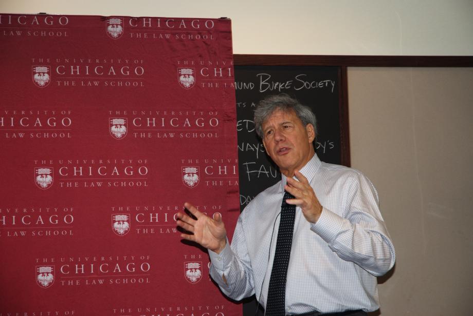 Stone, in a shirt and tie, motions with his hands in front of a step and repeat with the Law School logo.