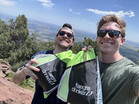 Two men in sunglasses hold up Faegre Drinker drawbags with a landscape behind them.