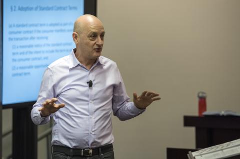 Omri Ben-Shahar gestures in front of a classroom with slides on a display screen behind him.