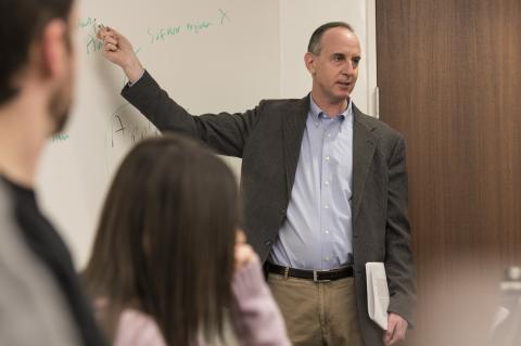 Eric Posner, in a blue shirt and grey sportscoat, points at the white board behind him.