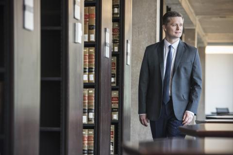 Anthony Casey, in a blue suit, white shirt, and blue tie, walks among the library stacks.