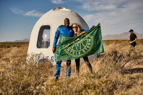 Wearing his blue jumpsuit, Jaison Robinson stands with a woman in dark clothes. They hold a flag. Behind them, the rocket nose sits in brush as a man walks toward it.