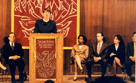 Janet Reno stands at a podium with seated students flanked on either side of her.