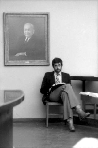 A man sits in a chair with his legs crossed one over the other with a large portrait hanging on the wall behind him.