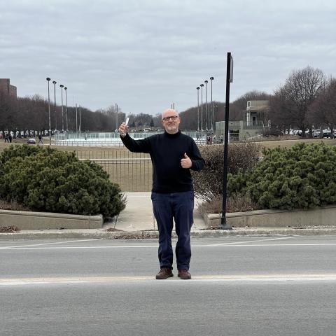 Professor Richard McAdams holds a champagne glass aloft and gives a thumbs up with his other hand as he stands in the middle of a road in the Midway.