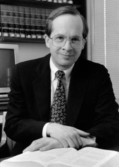 Black and white photograph of Douglas Baird with wire-frame glasses and in a suit and tie. A book is open in front of him.