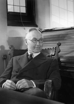 Black and white photograph of Harry Bigelow sitting at a desk in wire frame glasses and dark suit and tie.