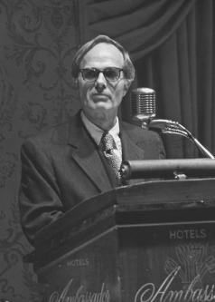 Black and white photograph of Phil Neal at a lectern in glasses and a suit and tie.