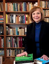 A woman in a black blazer and blue sweater stands in front of a bookcase.