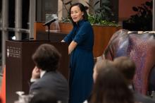 A woman stands at a lectern in front of a room of people listening her speak.