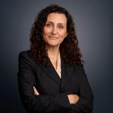 Professional headshot of Sandra Frantzen wearing a dark suit and beige blouse. Her hands are folded across her body.