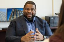 A man sitting at his desk gesturing while speaking to someone sitting across from him, not pictured.